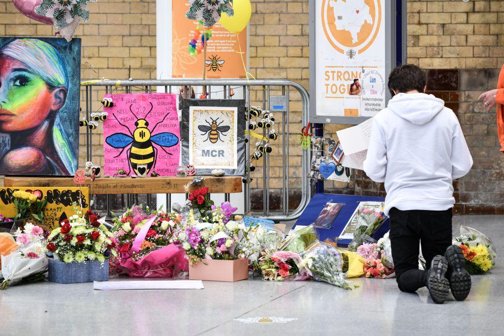 A young man kneels in front of the floral memorial at Victoria station during the commemoration. Colourful flowers, posters and pictures of Manchester bee icons are shown.