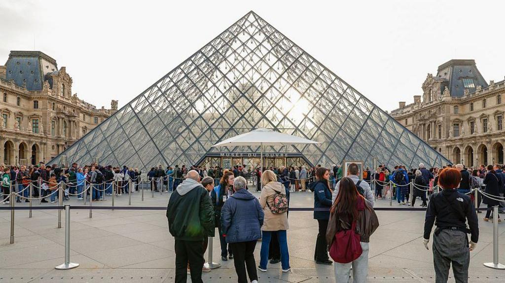 A queue of hundreds of people are lined up outside the Louvre's iconic pyramid-shaped glass entrance