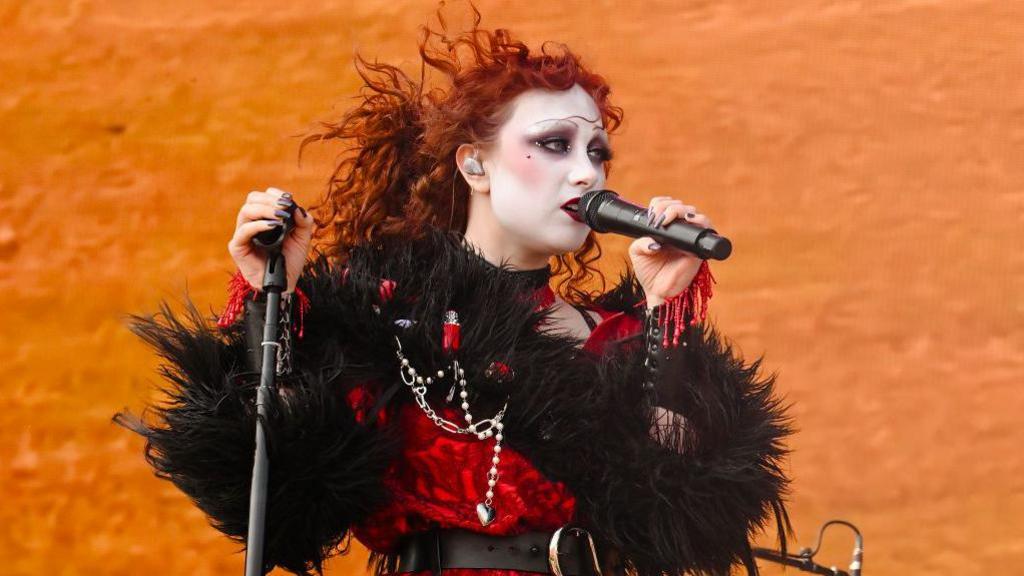 Chappell Roan performs during the 2024 Boston Calling Music Festival at Harvard Athletic Complex on May 26, 2024 in Boston. Chappell has bright red curly hair and wears a drag style make-up, complete with white face, strong blush and pencil thin brows. She wears a red and black feathered dress and sings into a mic in front of an orange backdrop. 