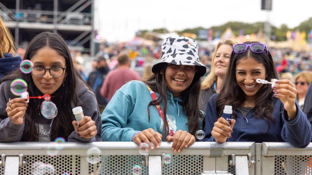 Three young women standing behind a barrier, with two on either side blowing bubbles from small tubs.