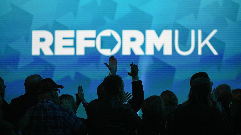 A Reform UK slogan projected on a wall with crowds pictured below in the dark.