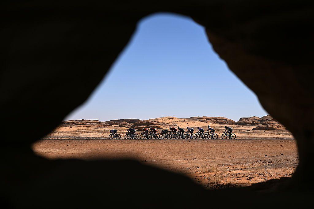 A line of cyclists rides through a vast desert landscape, seen through a natural rock opening, with red sand, distant cliffs, and clear blue sky framing the peloton’s endurance and isolation.