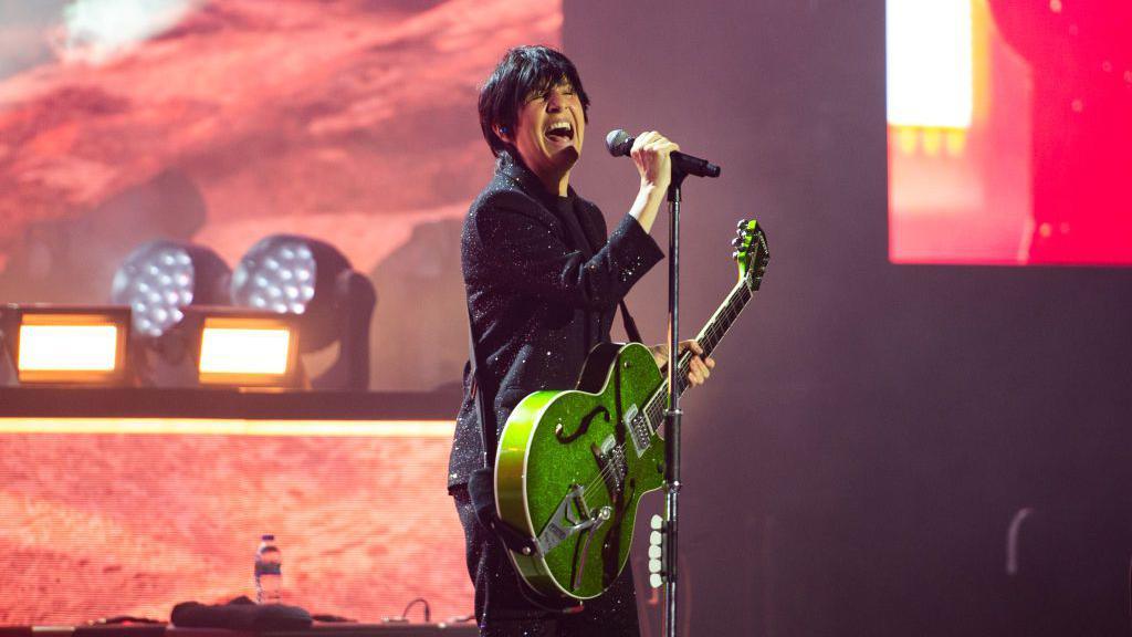 Sharleen Spiteri of Texas performs at The O2 Arena in London. She wears a black glittery suit and plays a green guitar. She is singing passionately. She has short cropped black hair.