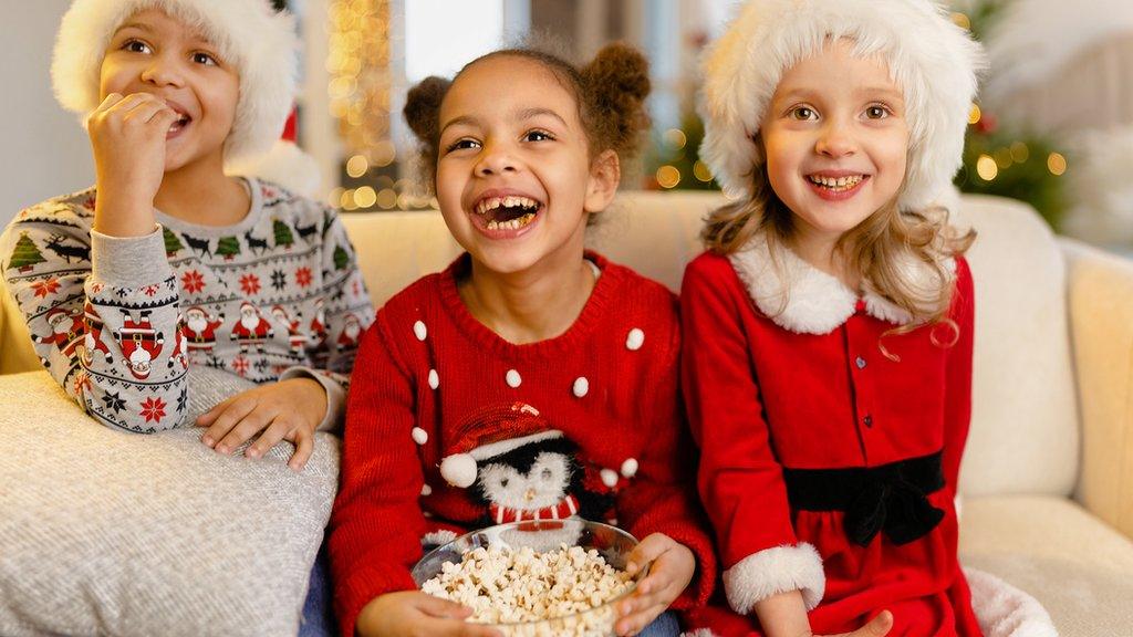 Three children in Christmas jumpers sit on sofa with popcorn