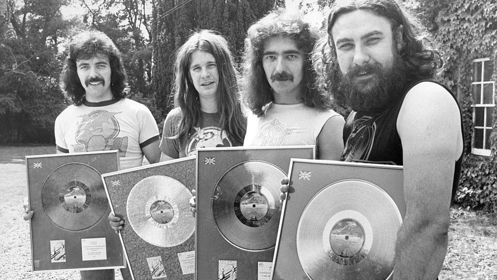 The four members of Black Sabbath looking young in a black and white photo. They are holding framed records and have long hair. 