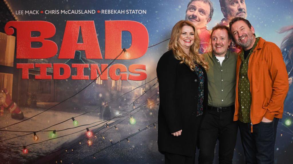 A woman in a black coat, a man in a green shirt and a man with a orange coat stand in front of a Christmas themed promotional banner. 