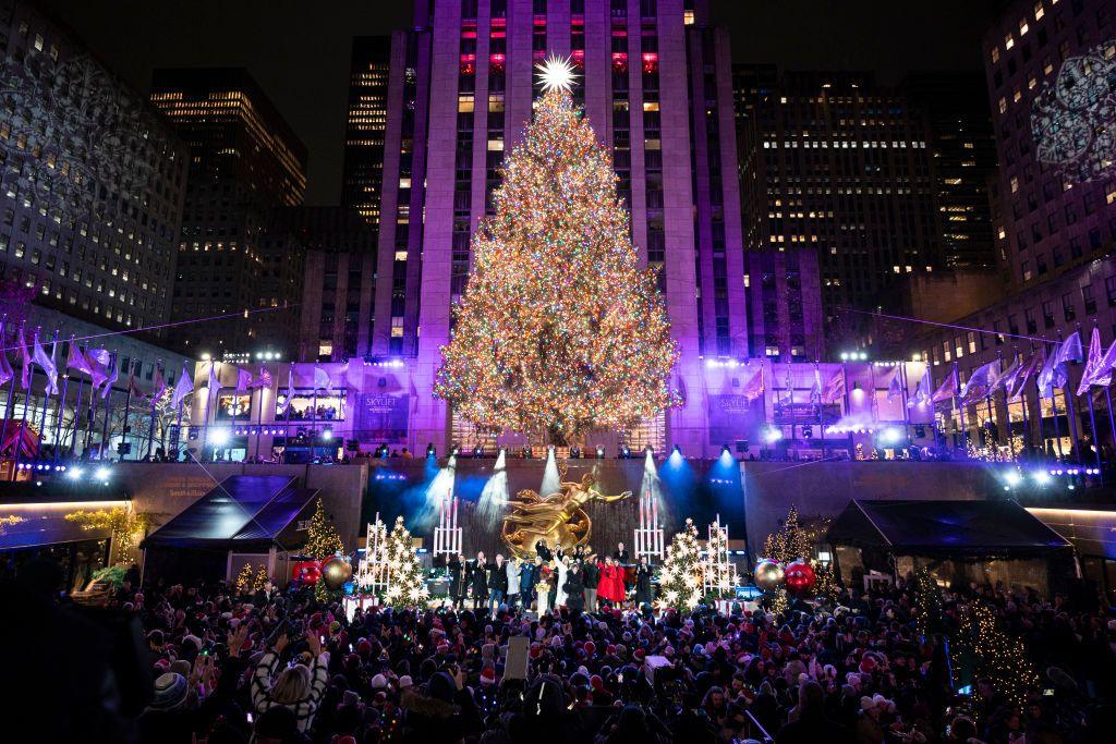 a bright and colourfully lit tree towers over a large group of people standing outside a big building while a small stage in front of the tree is filled with performers