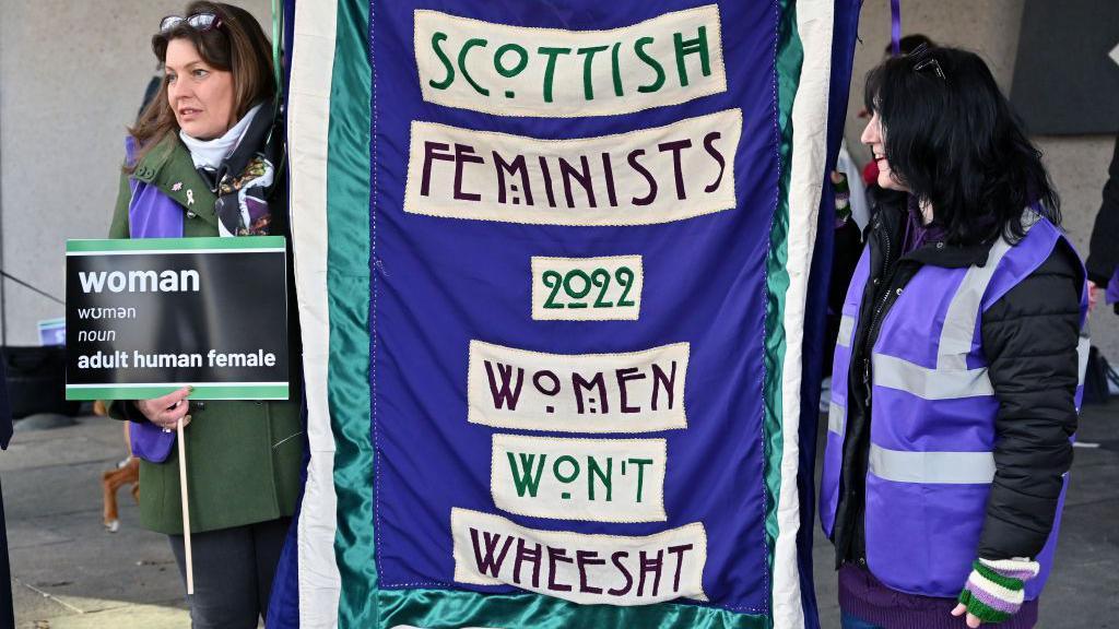 Two women holding up a purple banner with the words 'Scottish Femenists Women Won't Wheesht' on the front in white boxes.