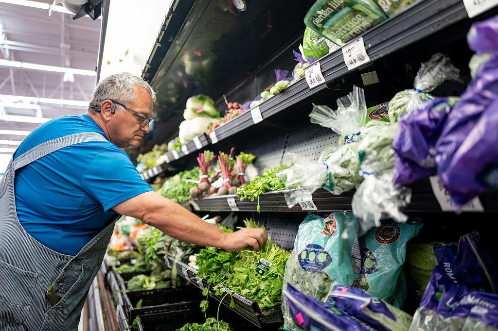 A man wearing a blue t-shirt and dungarees picks coriander from a fresh produce shelf in a supermarket.