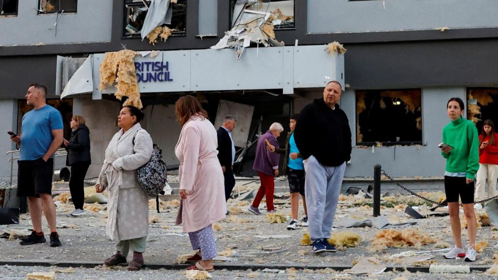 Ukrainians, some in their pyjamas and dressing gowns, stand outside a damaged building - there's debris on the floor