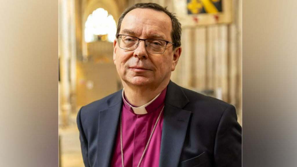The Right Reverend Philip Mounstephen is standing in a church, with the chain of a cross visible around his neck. He is wearing a suit and a burgundy shirt, with a white dog collar. He has glasses.