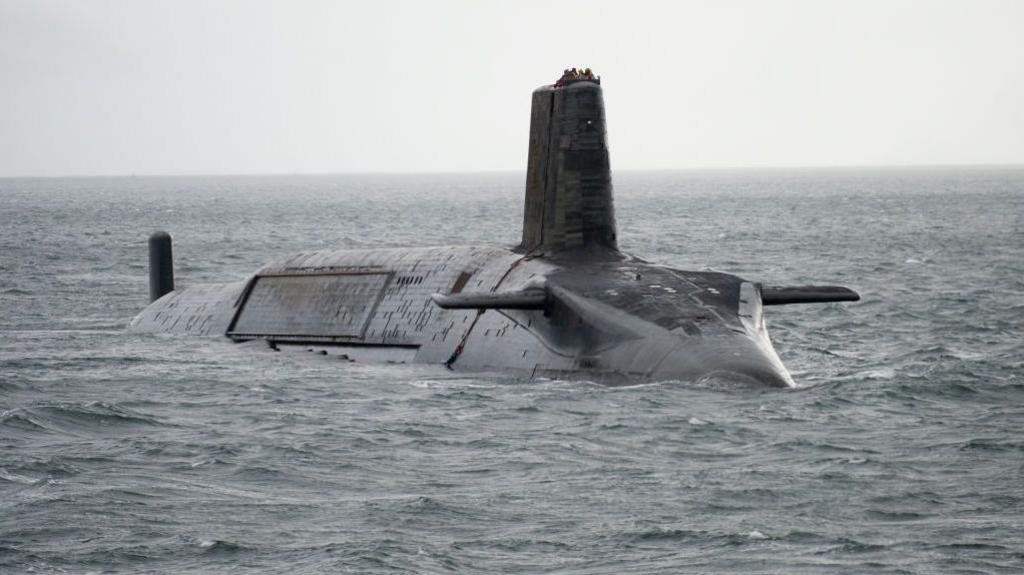 A large black Vanguard submarine with a group of sailors on its conning tower, is at sea. It is on the surface and it is a very grey day. The sea is slightly choppy.