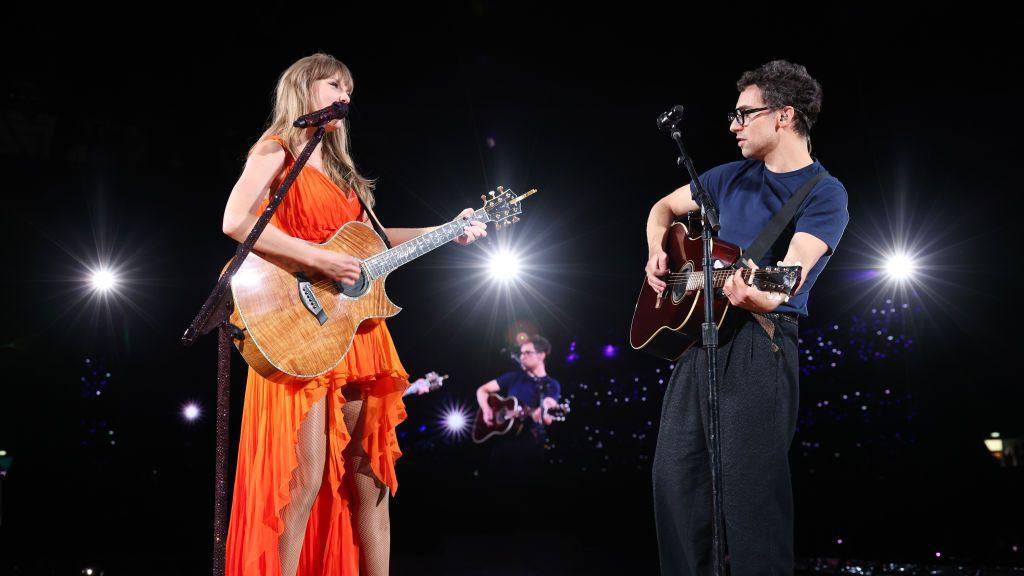 Taylor Swift plays an acoustic guitar with singer and producer Jack Antonoff, with lights illuminating them 