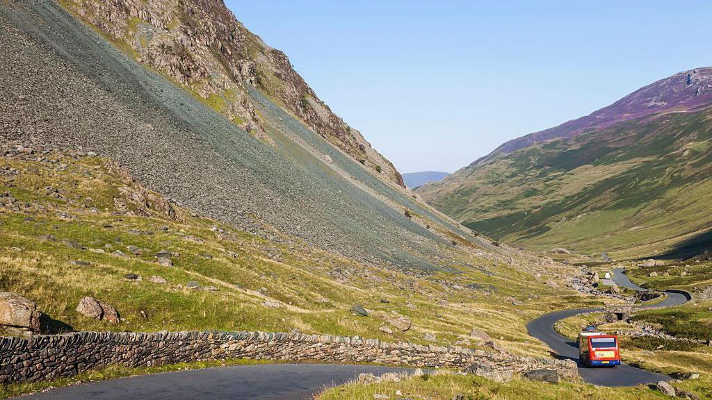 Honister Pass. A bus is being driven along a narrow road between two fells on a sunny day.