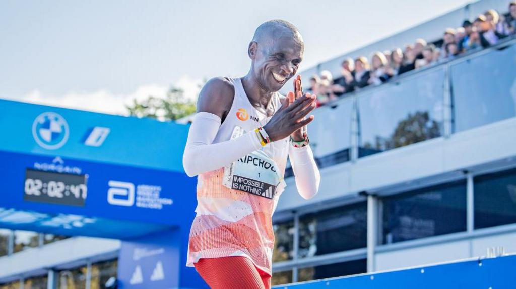 Eliud Kipchoge smiles after crossing the finish line to win the 2023 Berlin Marathon. The picture is taken from a low angle looking up at Kipchoge, who is wearing orange shorts and a white and orange running vest and white running sleeves. He also has a name bid on, with the phrase 'impossible is nothing' written at its base. Kipchoge is rubbing his hands together, while behind him is the blue finish gate with a digital clock across the top and a glass-fronted stand with a line of people watching from its roof
