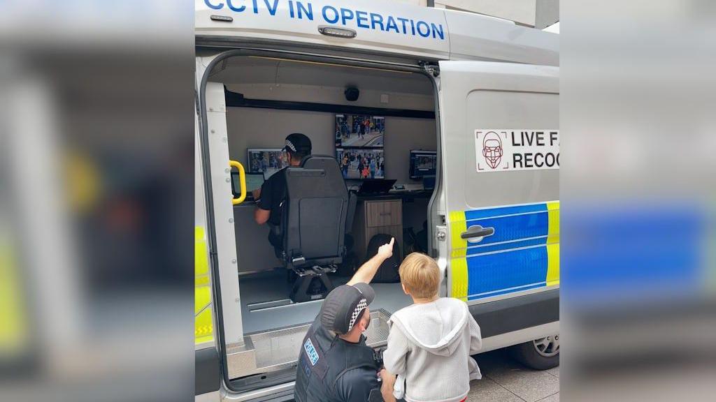 A police officer points inside a van as he shows a boy the technology behind the facial recognition cameras