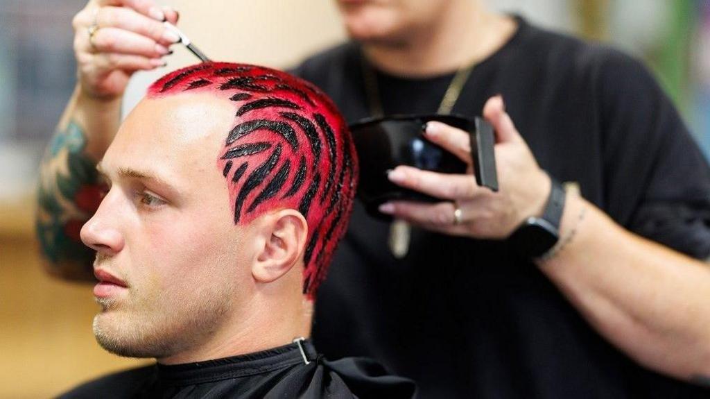 Bristol Bears rugby player Fitz Harding sits in a barber's chair. He is wearing the club's new red and black European kit and has had his hair dyed to match it.