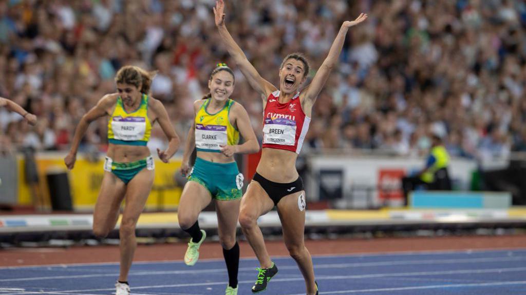 Runner Olivia Breen crosses the finish line of the 100m T38 race at the Commonwealth Games 2022, just in front of two other runners.
