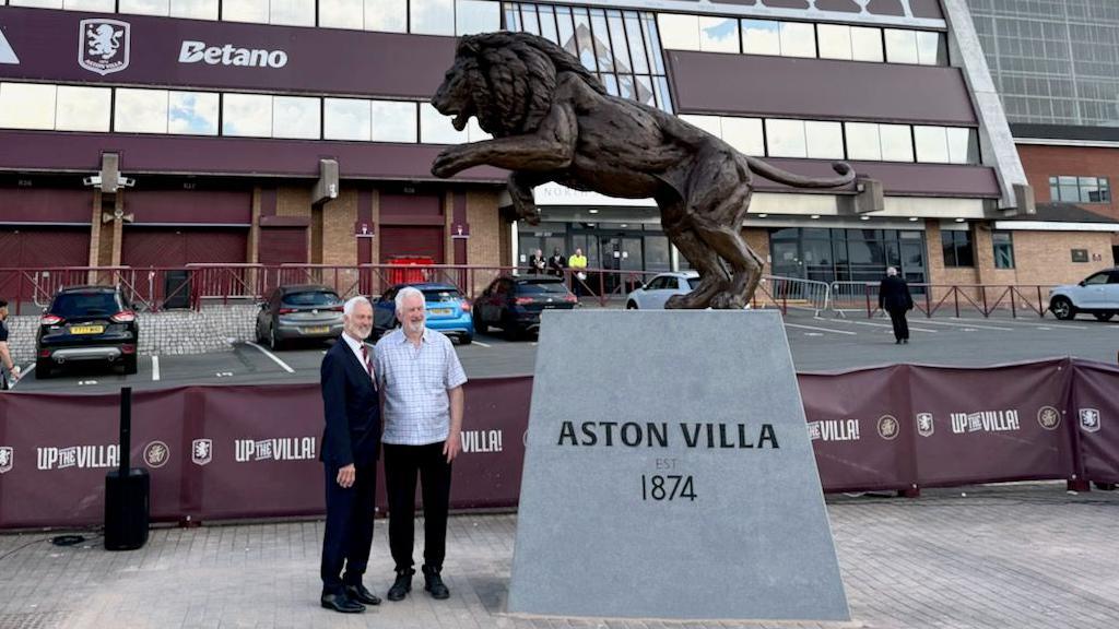 Two men standing next to a large, bronze lion statue behind the North Stand at Villa Park.