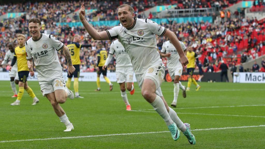 Michael Cheek celebrates after scoring for Bromley with his hand raised, running towards the corner flag at Wembley