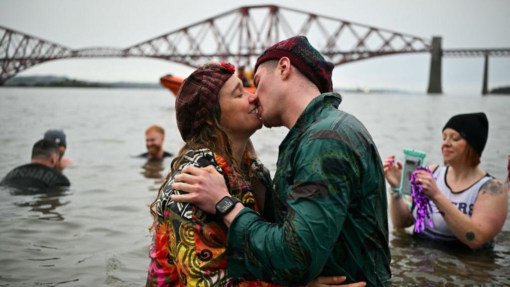 Particpants kiss during Loony Dook swim in Firth of Forth, near South Queensferry on New Year's Day. The man and woman are fully dressed and wearing hats, standing in the water in front of the Forth Rail Bridge. 