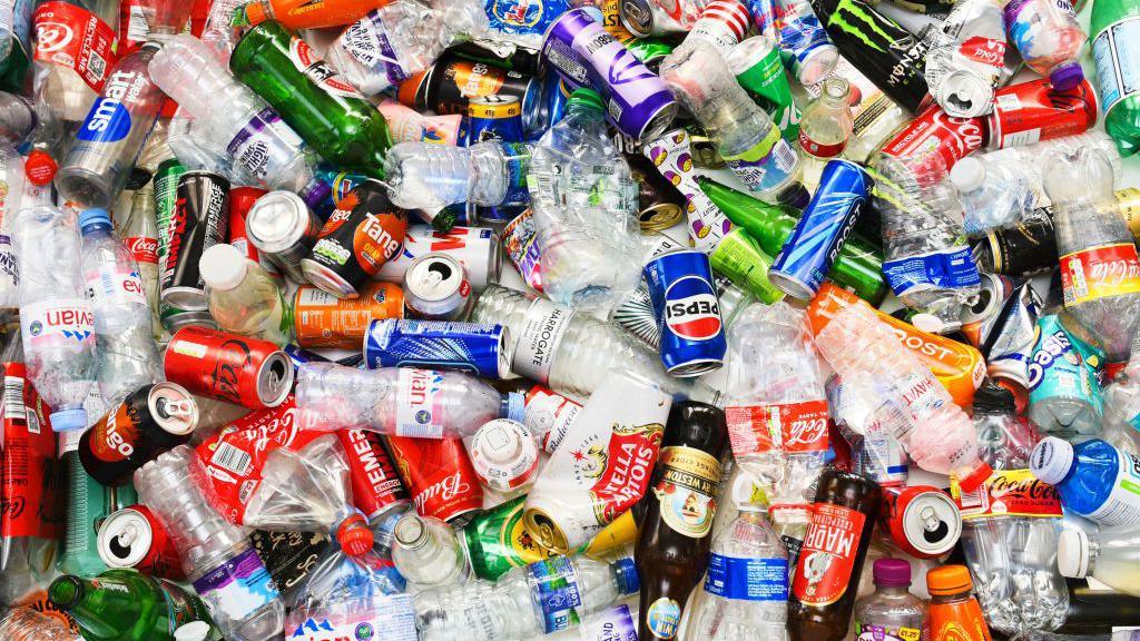An image of a mix of different drinks containers, all apparently empty and presumably awaiting recycling, including plastic water bottles, glass beer bottles, aluminium drinks cans and more, covering a variety of brands