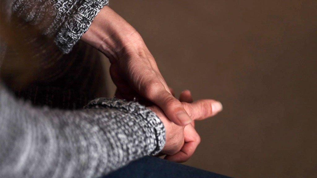 A blurred image of an unidentifiable woman sitting down with her hands clasped