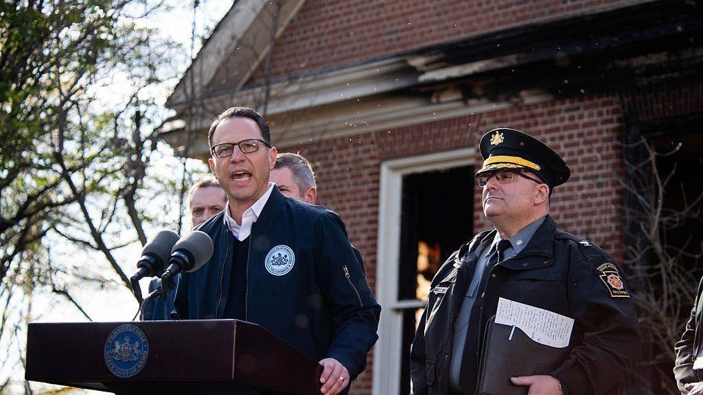 Governor Josh Shapiro speaks at a podium in front of a brick home with fire damage; he's wearing a jacket and glasses.
