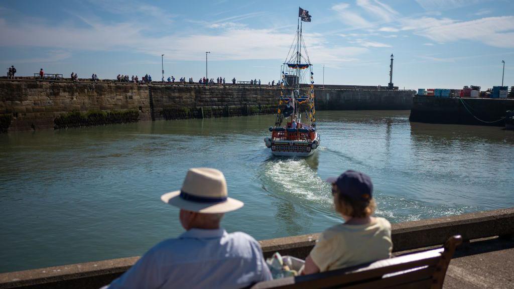 An elderly couple sat on a bench watch a pirate pleasure boat sailing out of Bridlington harbour.