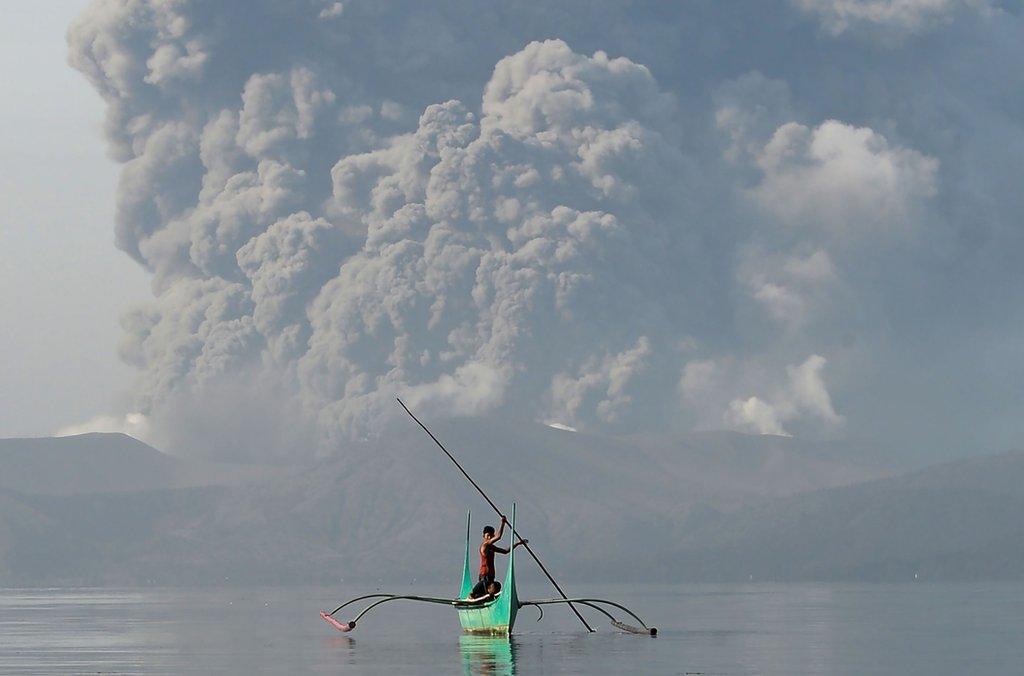 A youth living at the foot of Taal volcano rides an outrigger canoe while the volcano spews ash as seen from Tanauan town in Batangas province, south of Manila, on January 13