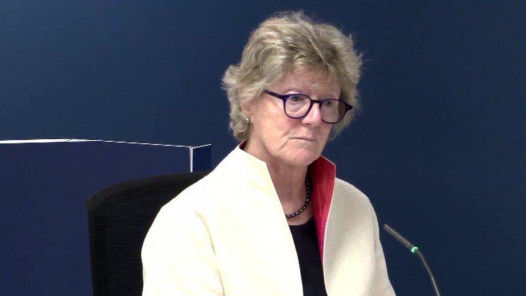 Sally wearing a white jacket and black rimmed glasses sitting in a chair with a microphone in front of her