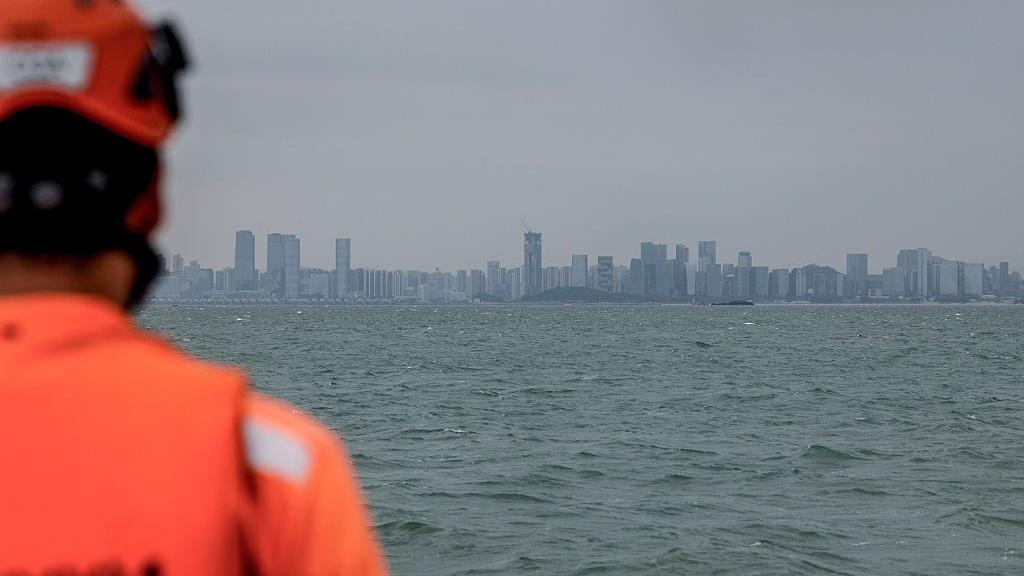 A Taiwanese coast guard in orange attire looks out to sea. In the distance is the a city skyline.