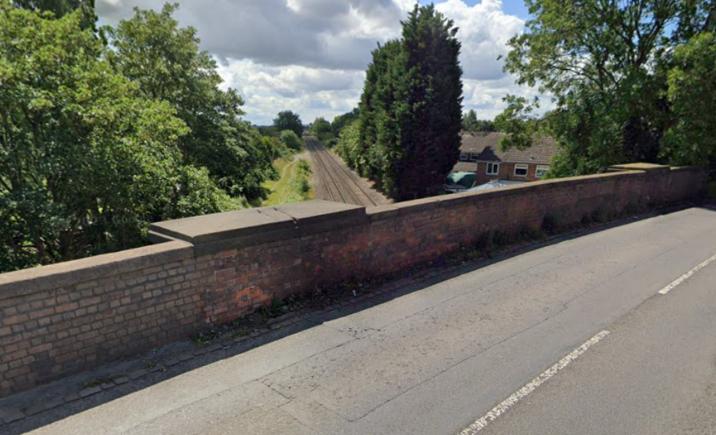 A view from a railway bridge with a road in the foreground, a brick wall and a railway line below it, with trees surrounding it