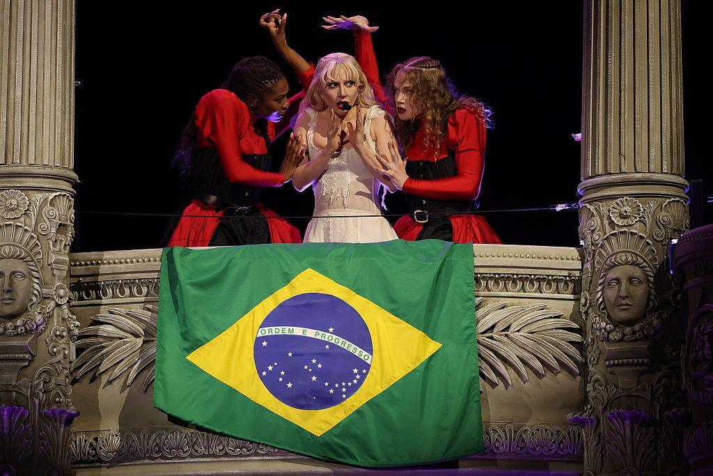 Lady Gaga in a white dress dances with two dancers dressed in red in front of a balcony draped with a Brazilian flag