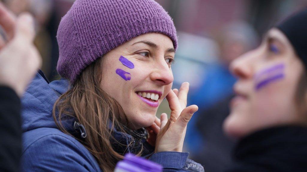 A woman wearing a purple hat puts purple face paint on her cheeks at march to mark International Women's Day