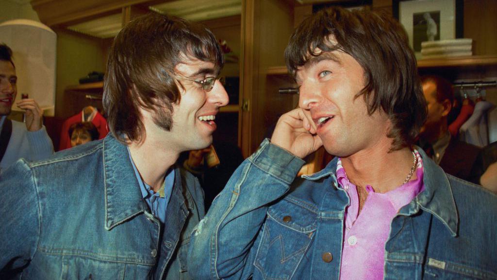 Liam and Noel Gallagher smiling while talking to each other. They are both wearing denim jackets and Liam is wearing a pair of glasses.