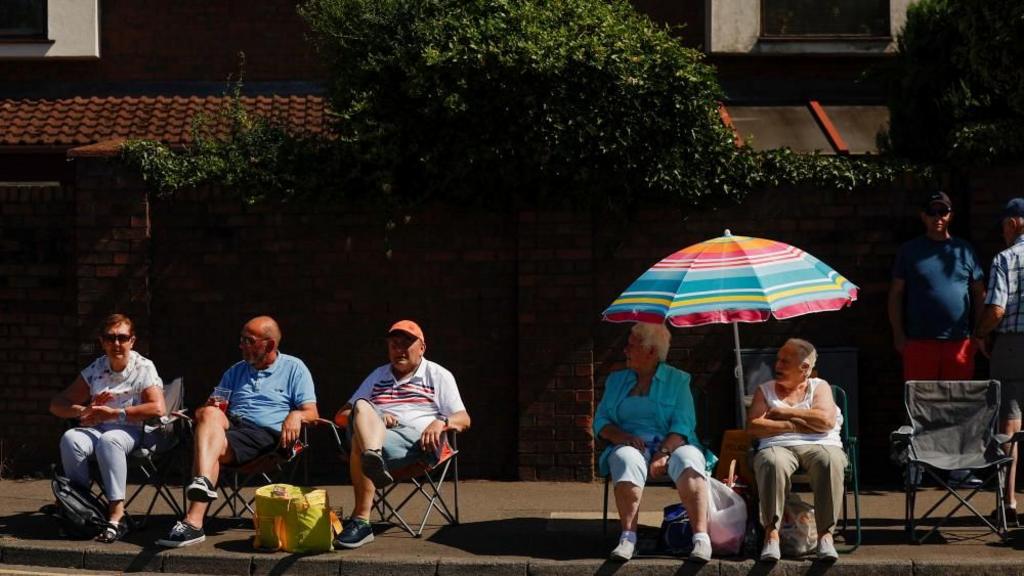 A group of elderly people sat on camping chairs on street, parasol above two women.