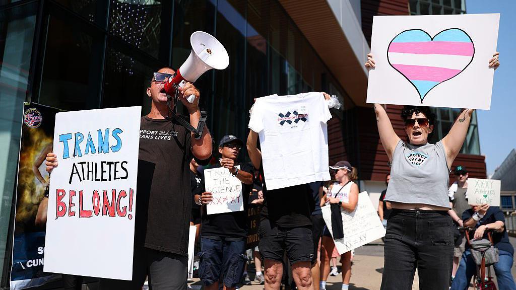Pro-trans protestors hold up signs while stood in front of anti-trans protestors