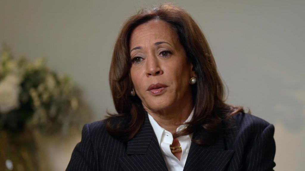 Close up of Kamala Harris in blue pinstripe blazer and white shirt, a clear rectangle chain ring necklace around her neck, a beige wall in the background