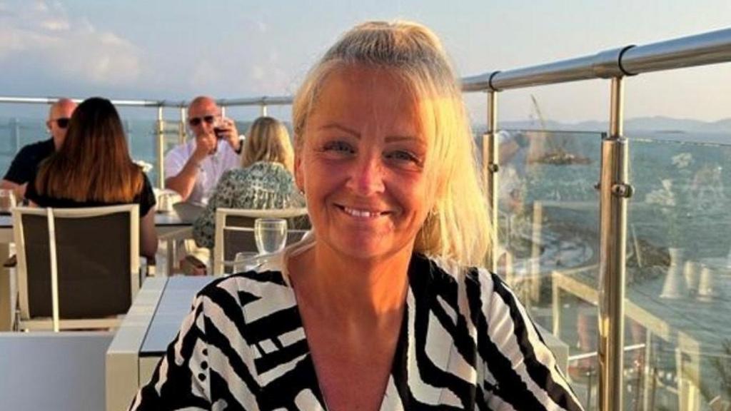 Anita Rose smiling sitting at a table on the roof of a tall building. She is wearing a black and white top. Other people are seating at a table behind her. There is a glass barrier next to her.