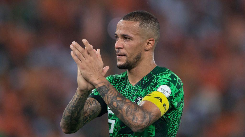 William Troost-Ekong wearing a Nigeria's green jersey and a yellow captain armband, looks sideways clapping