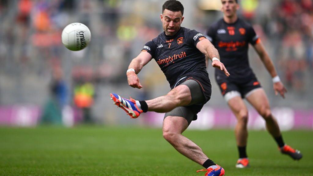 GAA footballer in a black kit kicking a white ball during a match. Another man of the same team looks on in the background