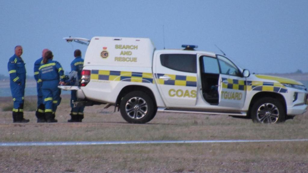 A white Coastguard vehicle with the words Search and Rescue and Coastguard written on the site. It is on a beach and beyond it is sea. There are four people in blue boiler suits at its rear door which is open.