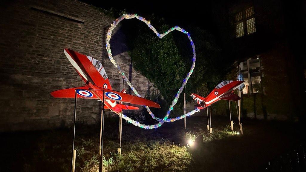A display at night, which has lighting from underneath and a wire in a heart shape with white tinsel and small coloured lights. On either end are two wooden red arrow planes fully painted red with RAF insignia.