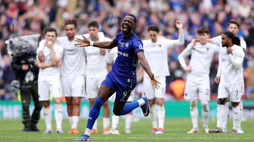 Rochdale's Mani Dieseruvwe runs celebrating with his arms extended, with dejected Boreham Wood players in the background 