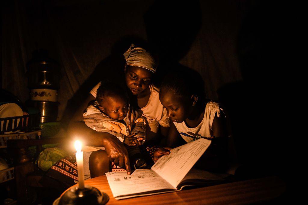 A young woman sits with a child and an infant at a table, reading from a school exercise book.