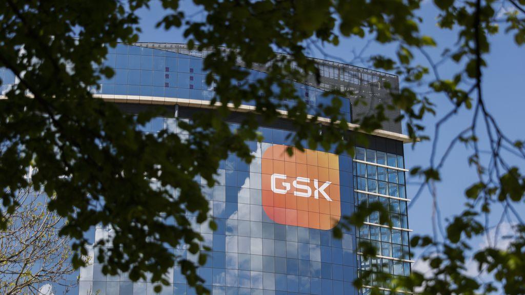 Picture of the GSK London headquaters. It is a tall, glass-fronted building, with a logo - an orange square containing GSK letters in white - at the top right. The sky is blue and there are tree branches in the foreground.