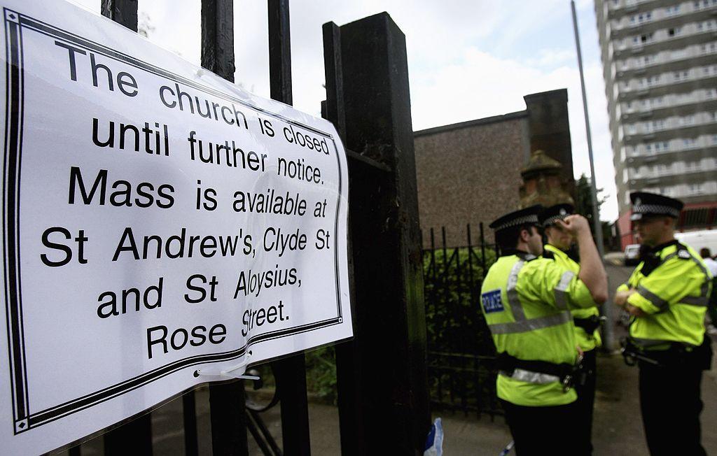 A sign on a fence reads "the church is closed until further notice" while three police officers stand off to the right. A high rise is visible in the background.
