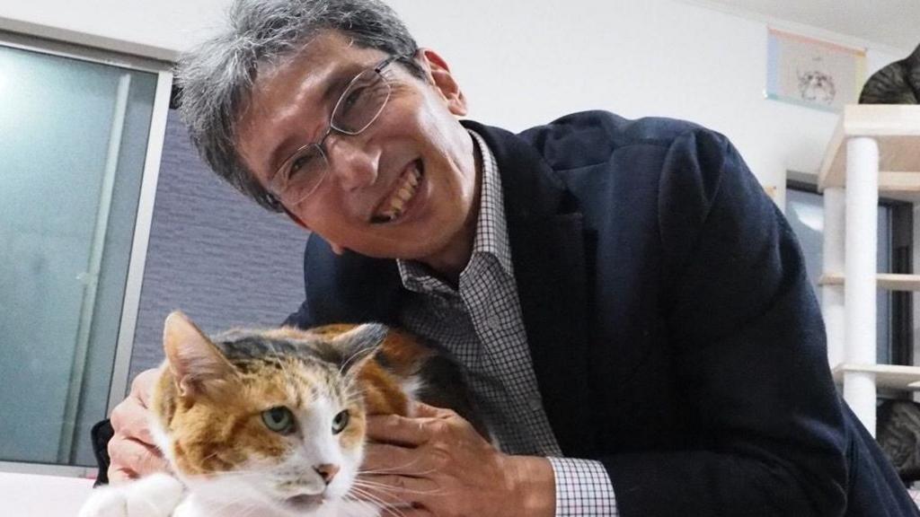Professor Sasaki smiles and leans down towards the table where he is holding a calico cat, mixed orange and brown fur with intently staring green eyes. They are in a cat rescue centre with other cats on a play tower in the background