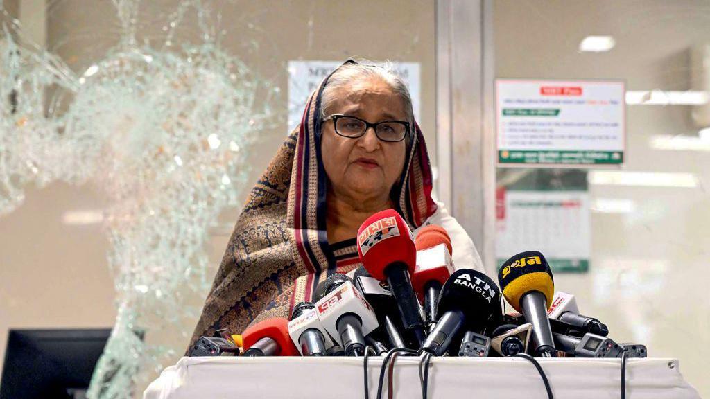 Sheikh Hasina, wearing a headscarf and glasses, speaks to reporters in front several microphones and with smashed glass behind her at a vandalised metro station in Mirpur on 25 July 2024.